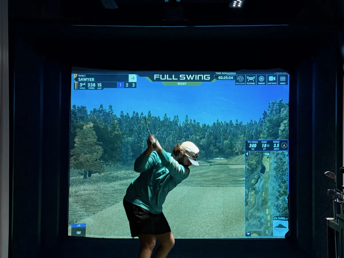 A golfer at the top of her backswing, a wooded fairway on the screen at Tee Off at The Wharf
