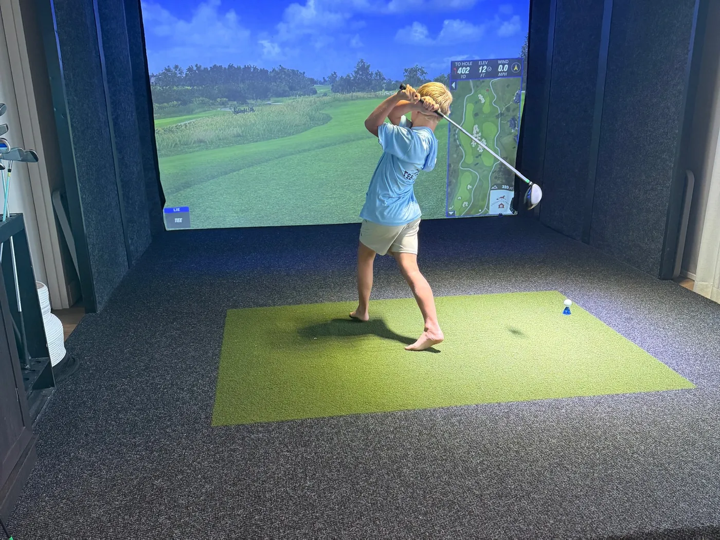 A golfer mid-swing on the turf of a bay at Tee Off at The Wharf, a fairway on the screen ahead