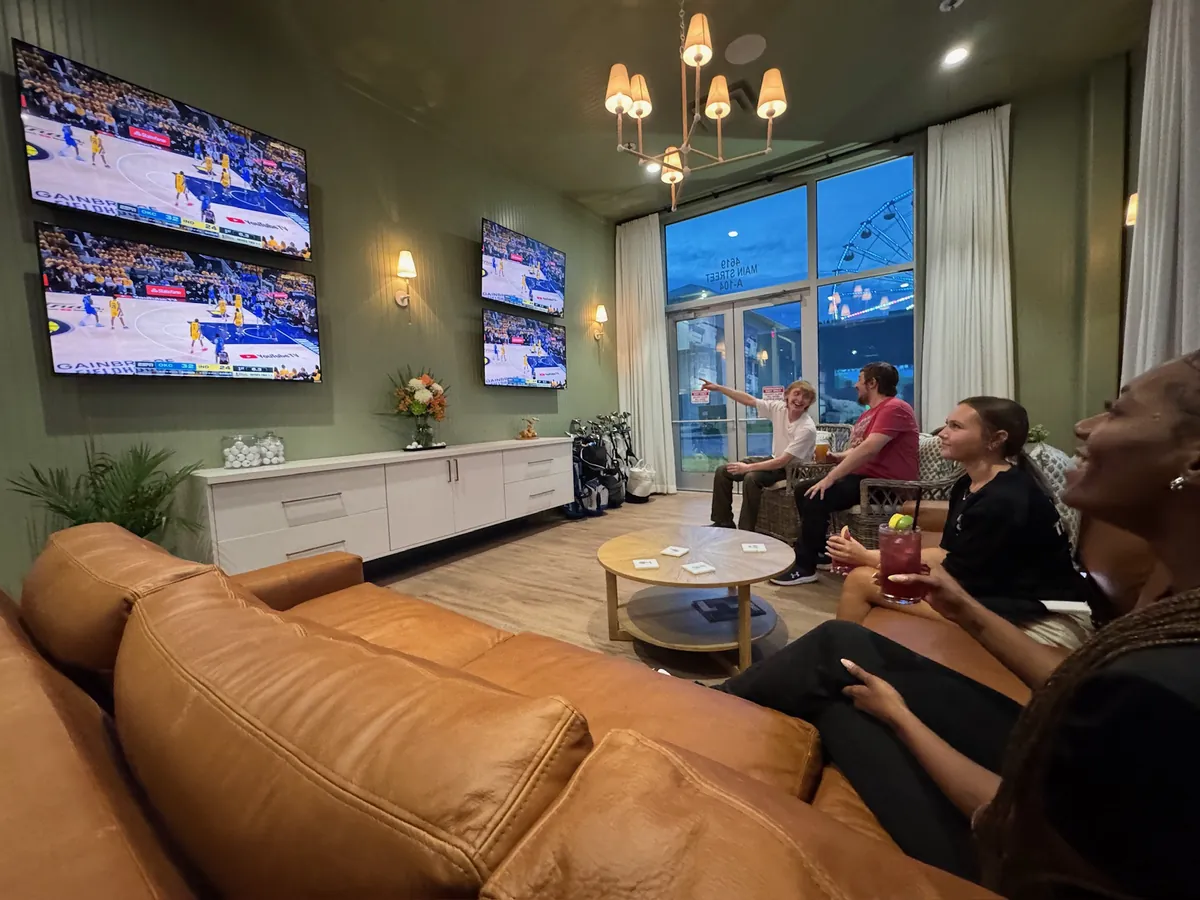 Guests on the leather sectional at Tee Off at The Wharf watching a basketball game on the wall screens, with the marina windows behind